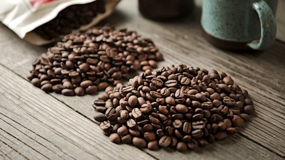 A wooden table featuring Robusta Coffee vs Arabica Coffee: dark brown, large Robusta beans and smaller, lighter brown Arabica beans. In the background, a mug and a bag of coffee beans are softly illuminated.