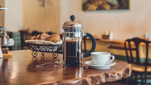 French press on a warm wooden countertop in a cozy café, with a cup, spoon, and basket of bread nearby; soft lighting and café décor in the background.